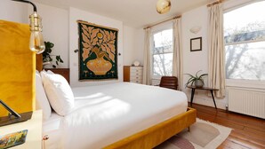 Quarto com cama, escrivaninha, cadeira, janela com cortinas, radiador e um quadro na parede.