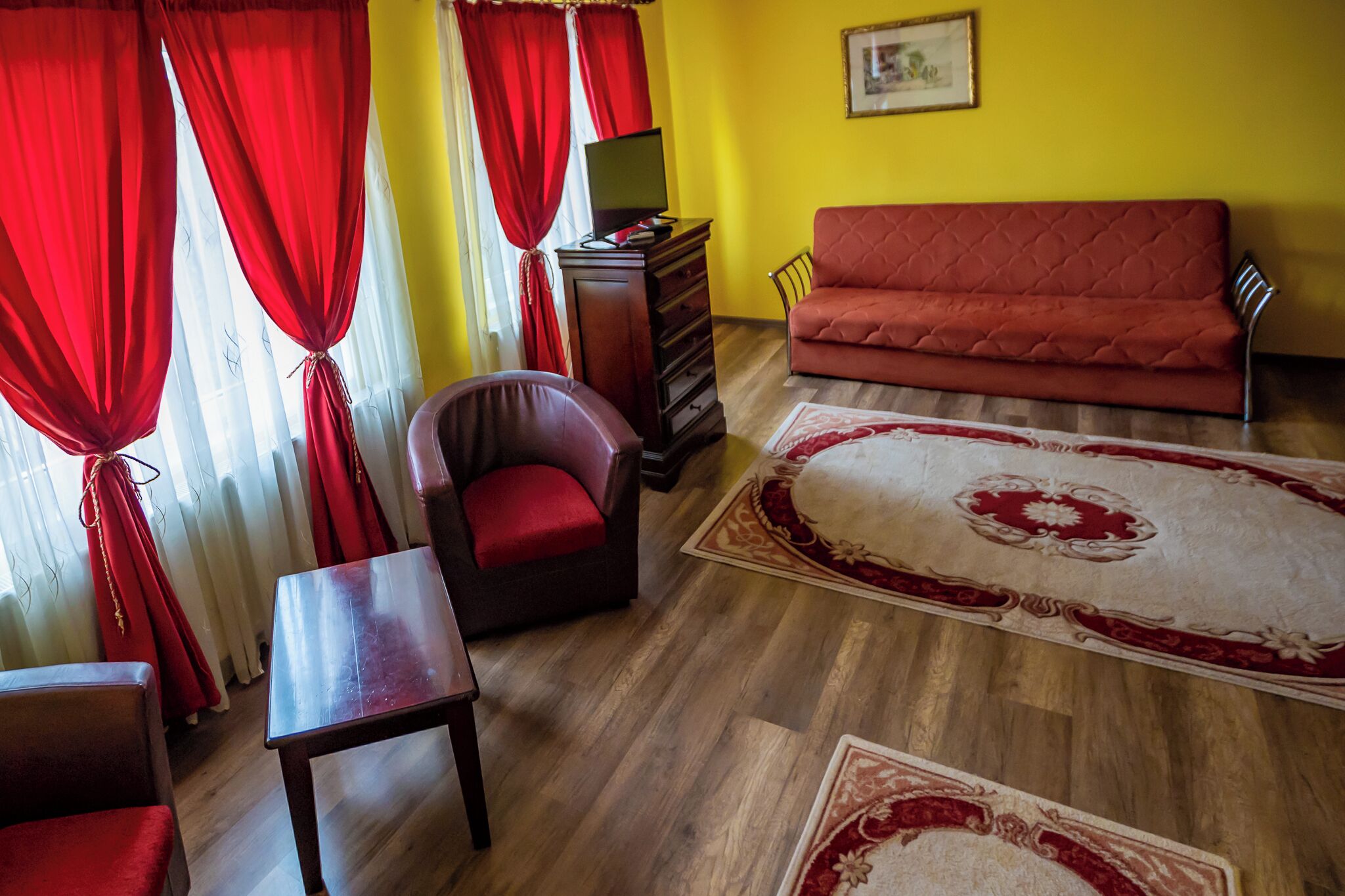Een kamer met een houten vloer, rode gordijnen, een rode bank, een fauteuil, een houten tafel en een televisie op een houten kast.