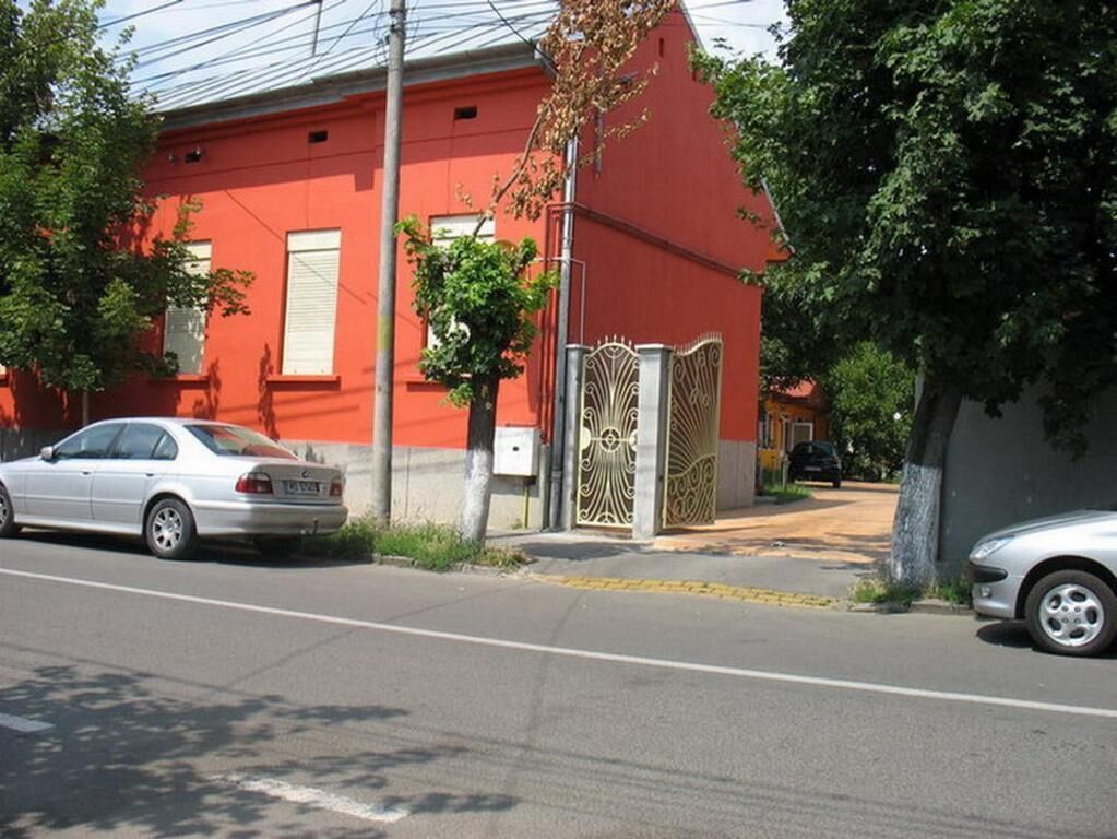 Un bâtiment rouge avec une entrée fermée par un portail, des voitures garées et des arbres.