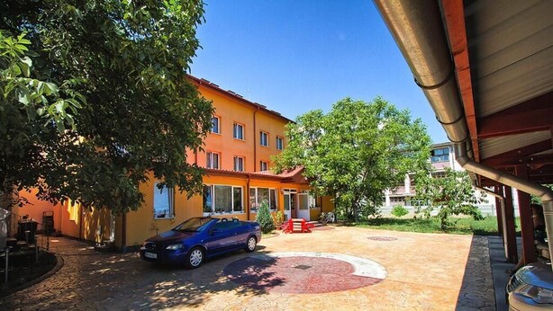 A courtyard with a blue car parked on a stone pavement, surrounded by trees and a yellow building with a red roof. - Noroc si Fericire (Târgu Mureș)