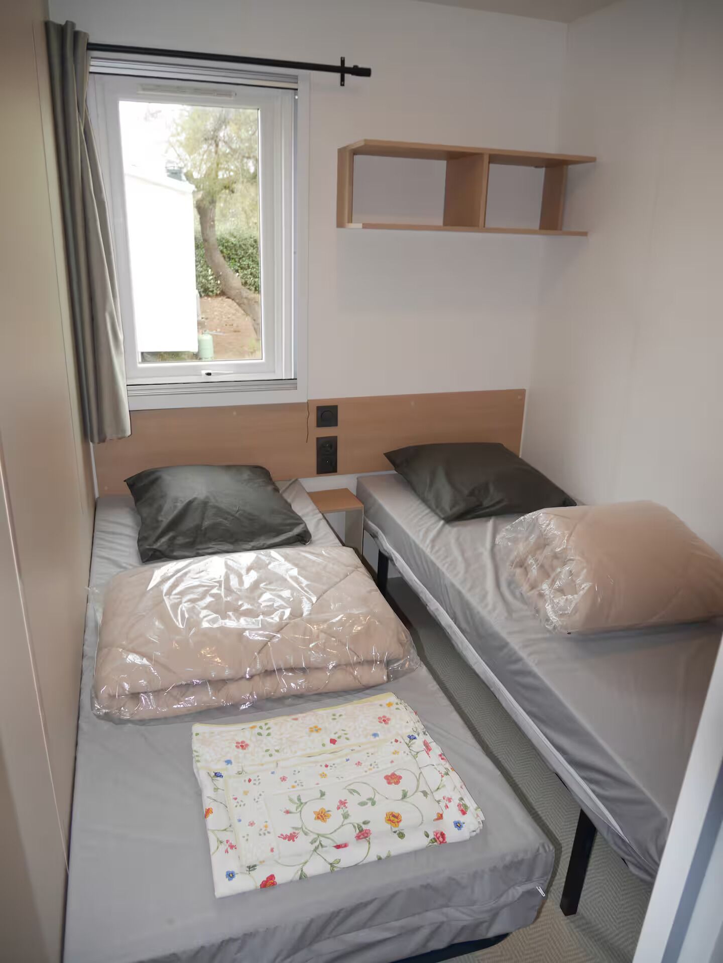 A small, compact room with two single beds, a window with curtains, a wooden shelf, and a floral bedspread.