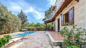 Eine terrasse mit Steinbelag, einem Schwimmbecken, Gartenmöbeln und einem Gebäude mit Holzläden.