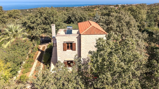 Ein Haus aus Stein mit rotem Ziegeldach, umgeben von üppiger Vegetation und mit Blick auf eine Küstenlandschaft.