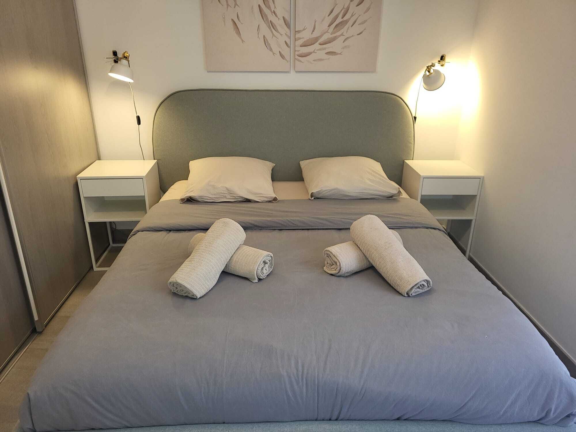 A neatly made bed with two pillows and two rolled towels, a bedside table, a wall-mounted lamp, and a framed artwork above the bed.