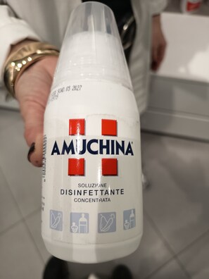 A person holding a bottle labeled 'AMUCHINA' with red squares and icons indicating it is a disinfectant concentrate. - VILLAGIOVEROOMS (Prato)