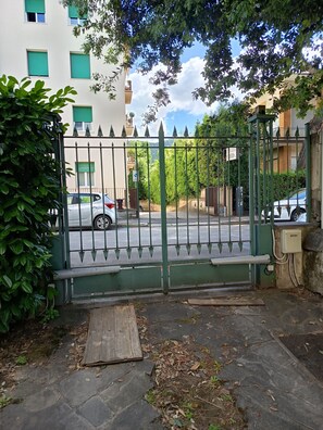 A gated entrance with a metal gate, a paved driveway, and a building with green shutters in the background. - VILLAGIOVEROOMS (Prato)