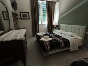 A bedroom with a bed, a bedside table, a mirror, a window with curtains, and a nightstand with a lamp. - VILLAGIOVEROOMS (Prato)