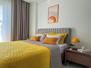 Un lit bien fait, avec une couette matelassée jaune et grise, une tête de lit rembourrée de couleur grise et deux coussins jaune moutarde.