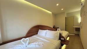 Habitación de hotel con dos camas, cabecero de madera, toallas dispuestas en forma de corazón, una mesita de noche y un baño pequeño con lavabo y espejo.