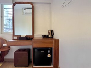 Quarto de hotel compacto com escrivaninha de madeira, pequena geladeira, espelho e janela com vista para edifícios.