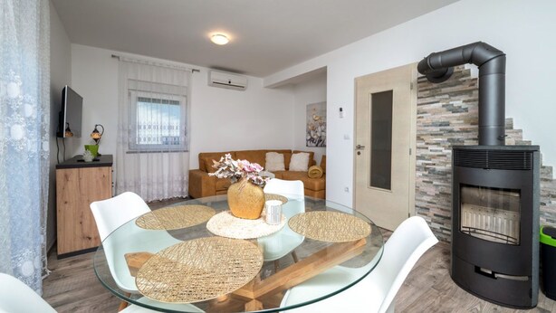 A modern living room with a glass dining table, white chairs, a sofa, and a stone fireplace. - Apartman Sabina 2 by Villas Guide (Fažana)
