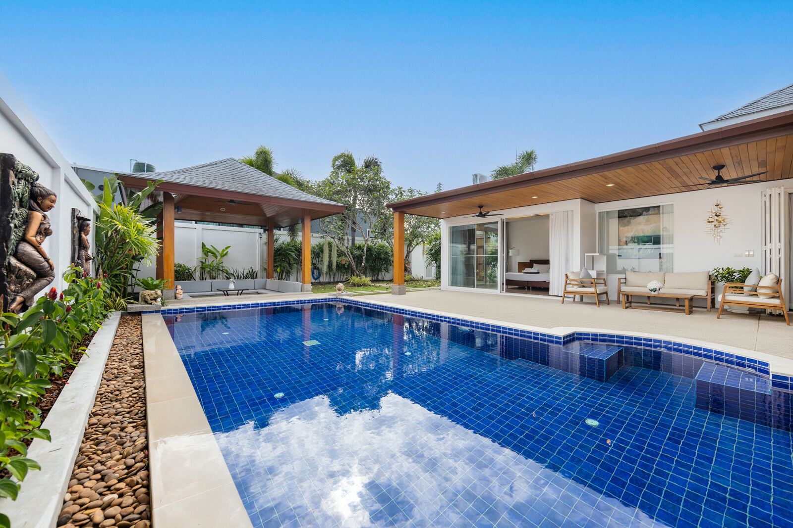 A swimming pool with a tiled deck, a covered seating area, and a modern house with a patio.
