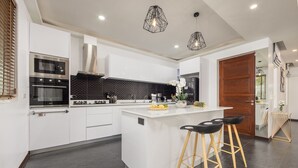 A modern kitchen with white cabinetry, a central island, black countertops, and wooden stools. - Villa Hemi 2BR Modern Thai Style (Choeng Thale)