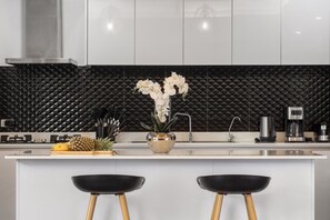A modern kitchen with a black textured backsplash, stainless steel appliances, and a central island with stools. - Villa Hemi 2BR Modern Thai Style (Choeng Thale)