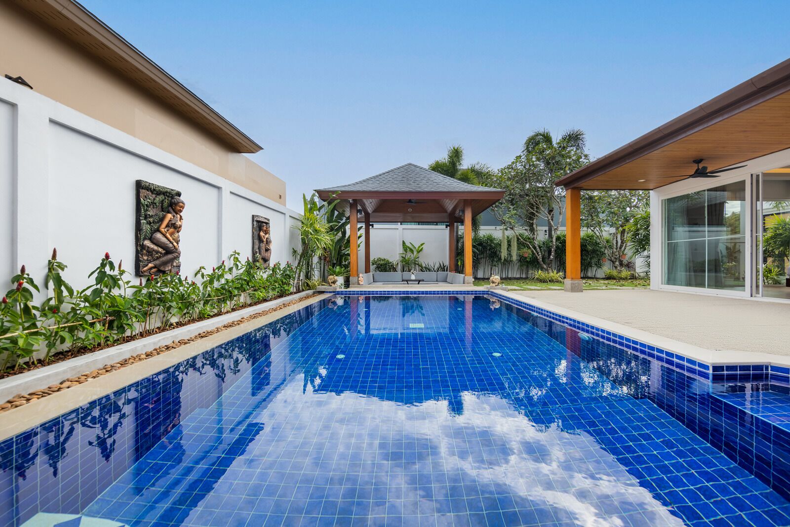 A swimming pool with a tiled deck, surrounded by plants and a covered seating area.