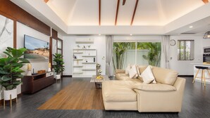 A modern living room with a sectional sofa, a flat-screen TV, a wooden coffee table, and a built-in bookshelf. - Villa Hemi 2BR Modern Thai Style (Choeng Thale)
