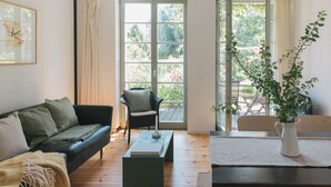 A living room with a sofa, a chair, a coffee table, and a dining table. There are large windows and a view of greenery outside. - Turmvilla Werder - Historic vacation home with garden, Havel view & bathing jetty (Werder Havel)