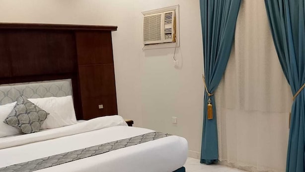 A bedroom with a bed, a headboard, a window with curtains, and a wall-mounted air conditioning unit. - Assistanceforfurnishedresidential units (Jeddah)