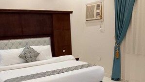 A neatly made bed with a headboard, a bedside table, and a window with a curtain. - Assistanceforfurnishedresidential units (Jeddah)