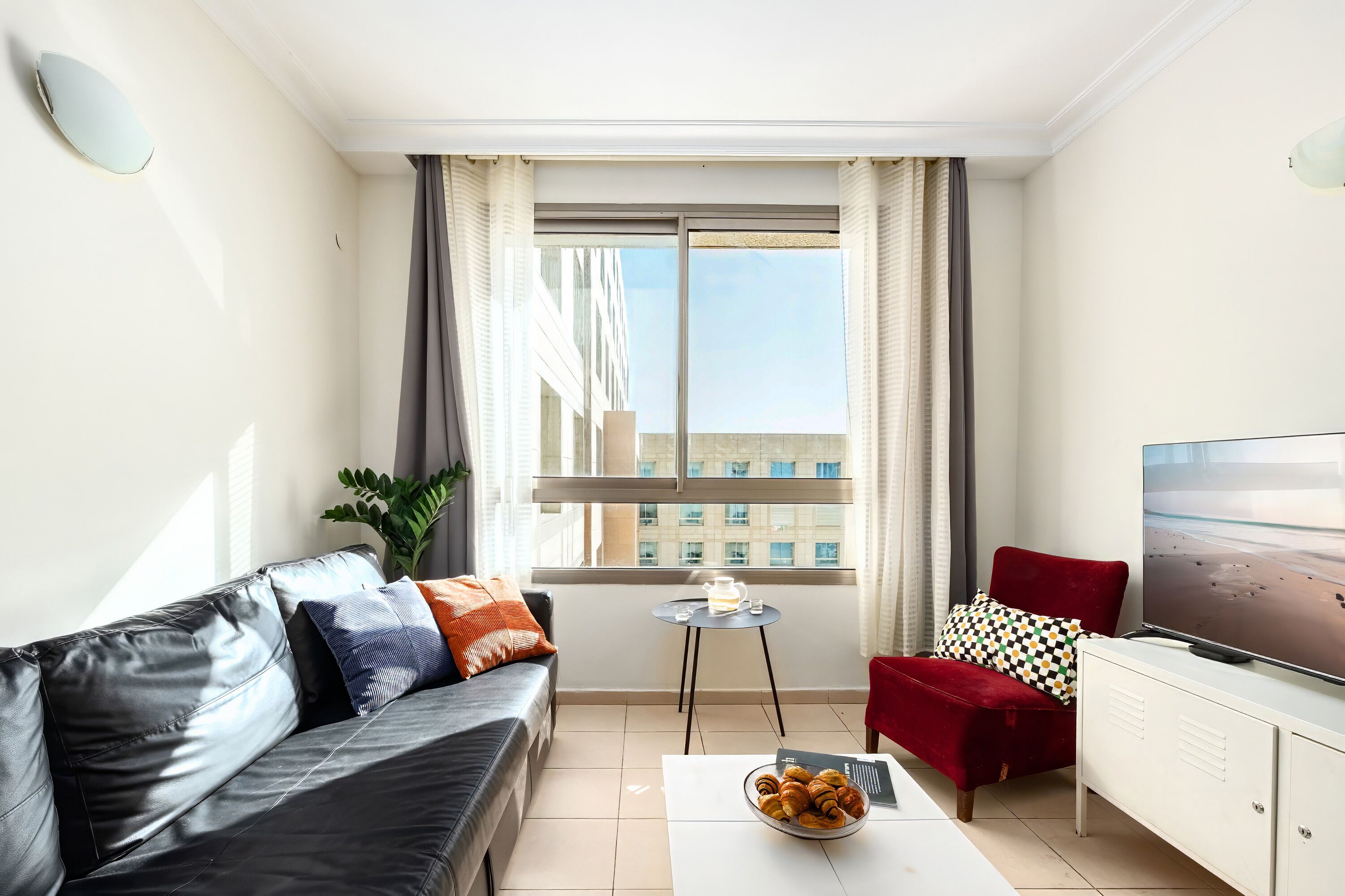 A modern living room with a leather sofa, a red armchair, a TV, a small table with a bowl of pastries, and a large window with a city view.
