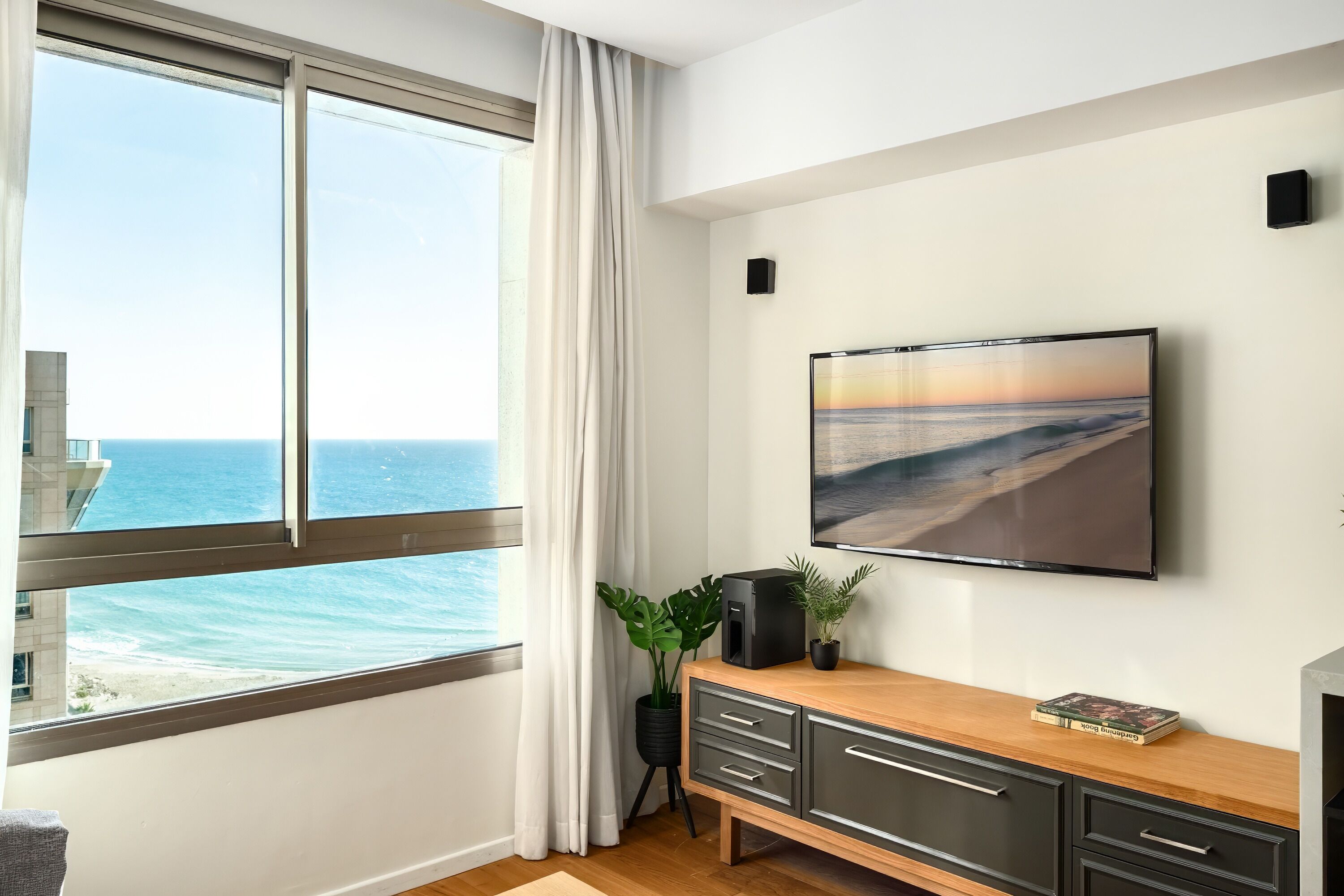 A modern living room with a large window overlooking the ocean, a flat-screen TV mounted on the wall, and a wooden cabinet with drawers.