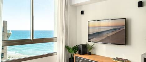 A modern living room with a large window overlooking the ocean, a flat-screen TV mounted on the wall, and a wooden cabinet with drawers.