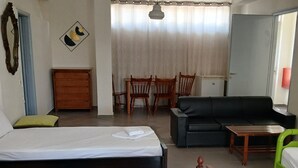 A hotel room with two beds, a sofa, a dining table with chairs, a mirror, and a window with curtains. - GOLDEN BEACH (Aegina)