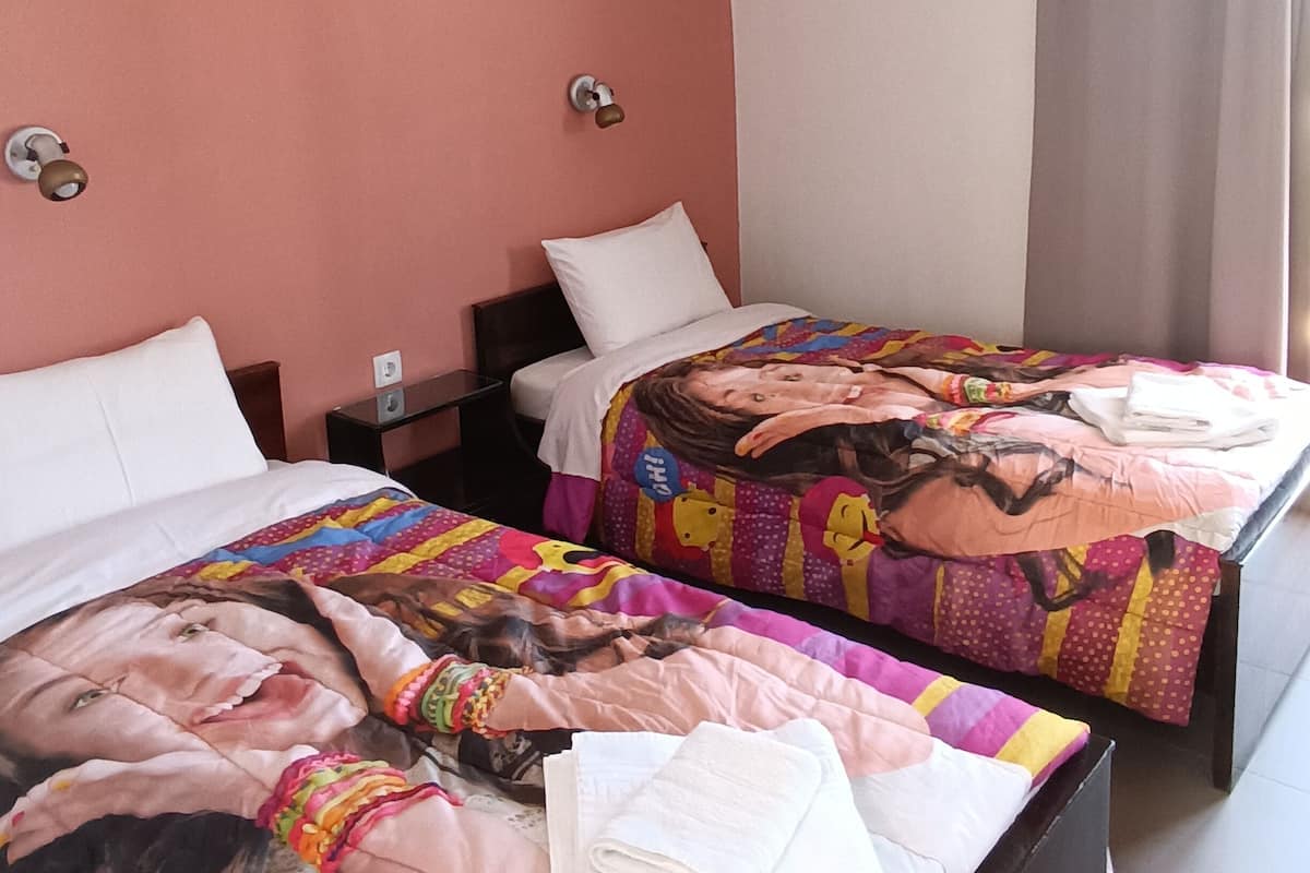 Habitación de hotel con dos camas, cada una con un edredón colorido y almohadas blancas. Entre las camas hay una mesita de noche con una lámpara, y una ventana con cortinas.