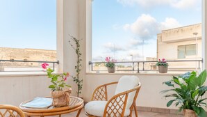 Een balkon met rieten meubilair, planten in potten en uitzicht op gebouwen.