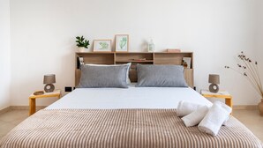 Een netjes ingerichte slaapkamer met een bed, kussens, een nachtkastje, een lamp, een plank met boeken en decoratieve objecten, en een vaas met gedroogde takken.