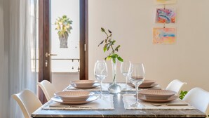 Een eettafel gedekt met borden, glazen en een vaas met planten, met kunst aan de muur en uitzicht op een balkon met beplanting.