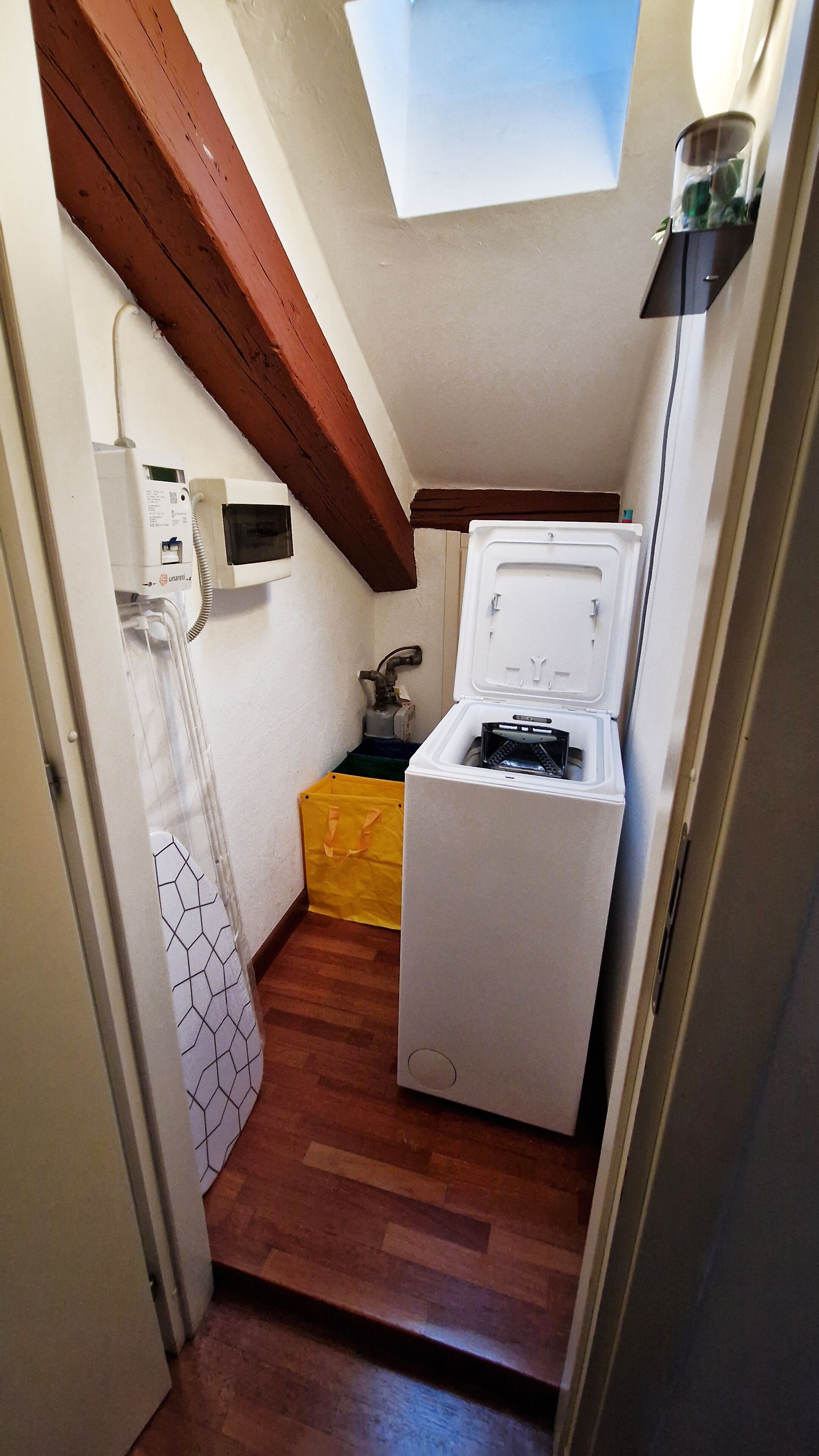 A small, narrow room with a white washing machine, a yellow laundry basket, and a skylight.