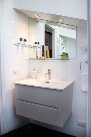 Une salle de bain moderne avec un meuble-lavabo blanc, une étagère en verre où sont rangés les articles de toilette et un grand miroir au-dessus du lavabo.