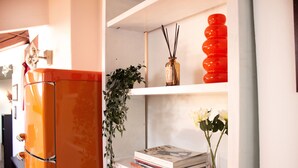 Een keuken met een moderne oranje koelkast, een witte plank met boeken en een bloevas, en een rode decoratie.
