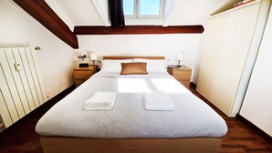 Een netjes opgemaakt bed met witte lakens en een bruin kussen, een nachtkastje met een lamp, en een bijzettafeltje met een klok.