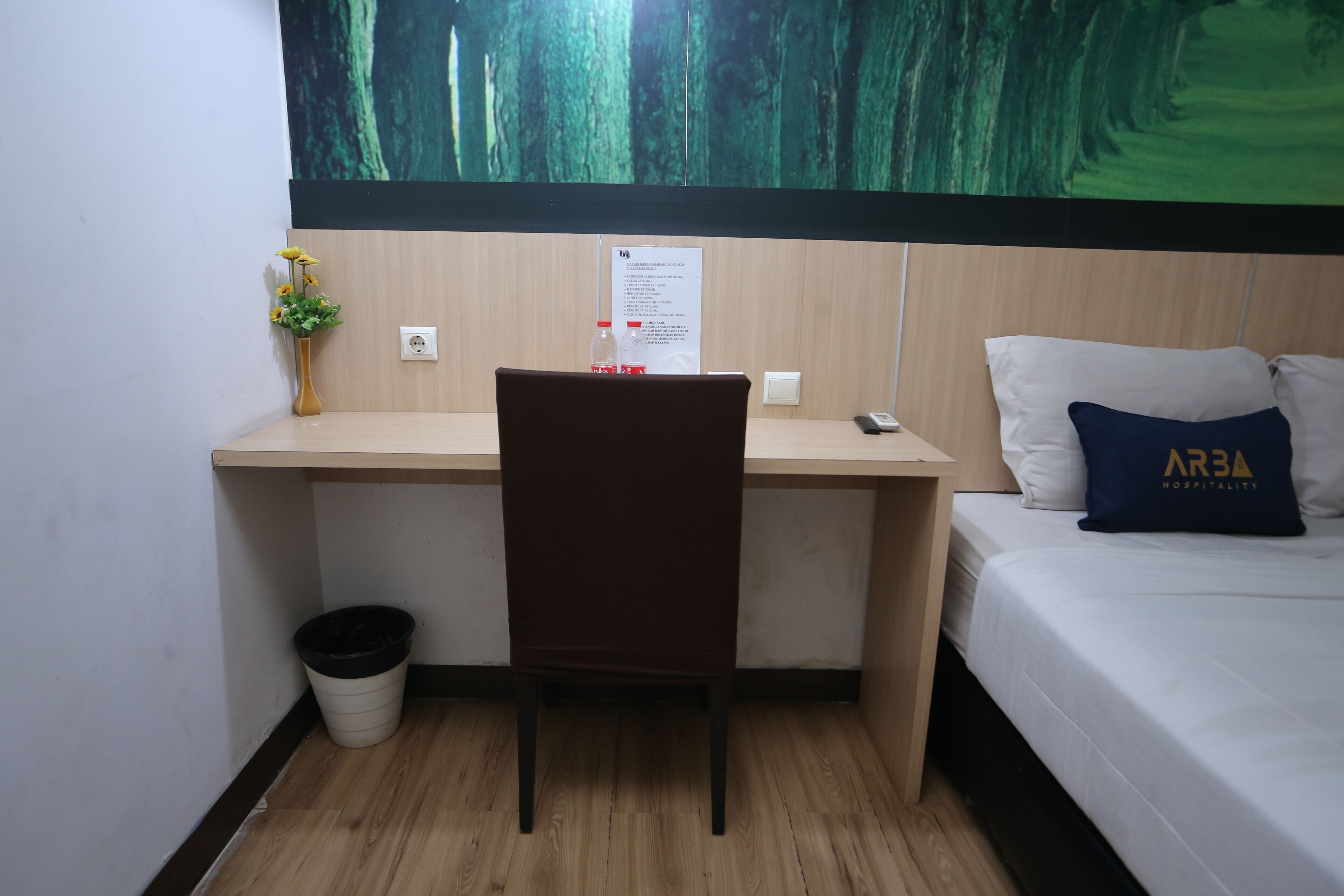 Hotellihuone, jossa on puinen työpöytä, tuoli, valkoisilla lakanoiden ja tyynynpäällisillä varustettu sänky, jossa on yrityksen »ARBA Hospitality» -logo, pieni kasvi työpöydällä ja roskakori.