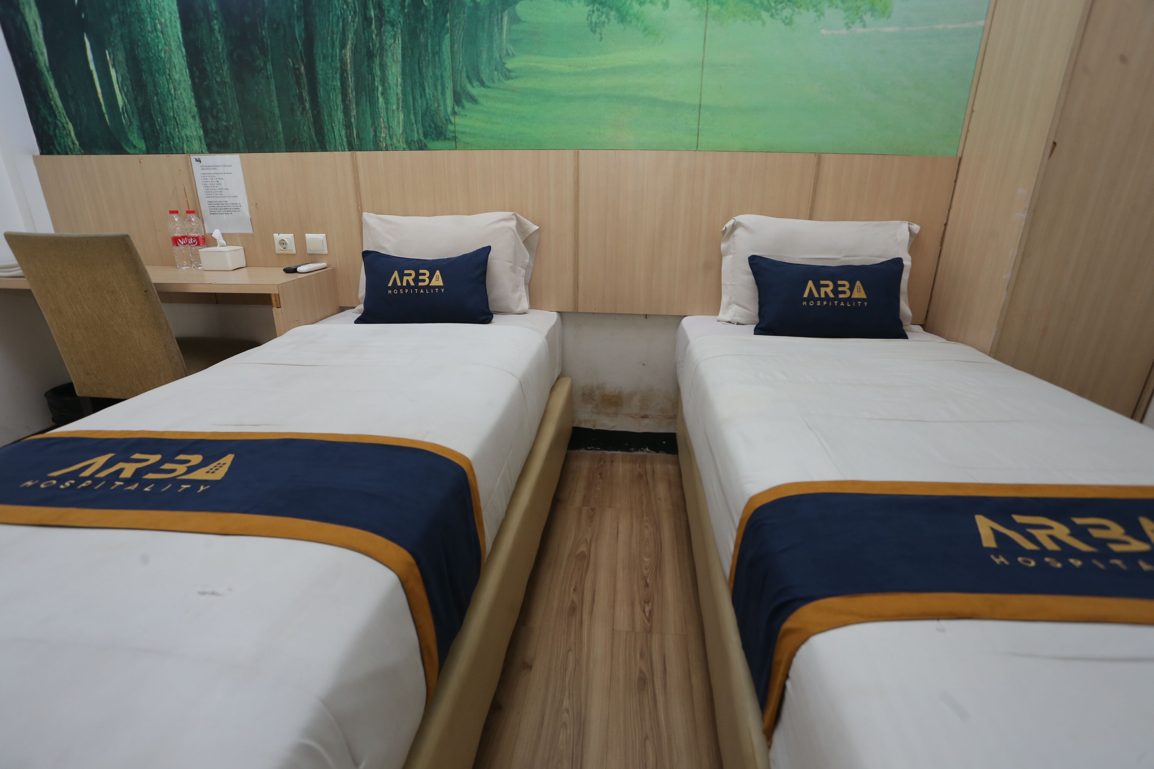 A hotel room with two beds, each with a blue and white bedspread featuring the ARBA Hospitality logo. There is a wooden desk with a chair, a small table, and a bottle of Coca-Cola. A poster of a golf course is on the wall behind the beds.