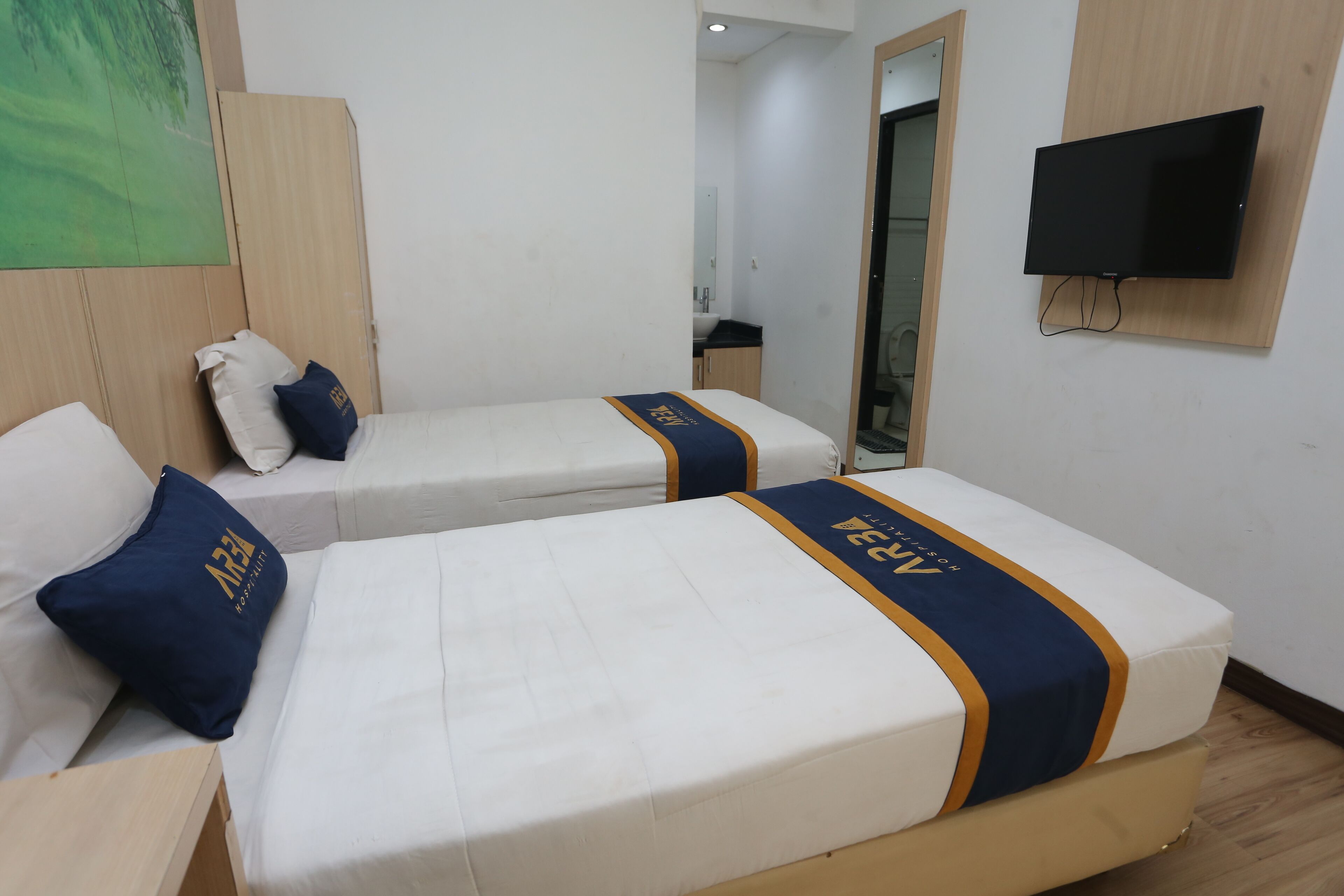 Ein Hotelzimmer mit zwei Einzelbetten, jeweils mit blauem und goldenem Bettüberwurf, weißer Bettwäsche und passenden Kissen. Ein Flachbildfernseher ist an der Wand befestigt.