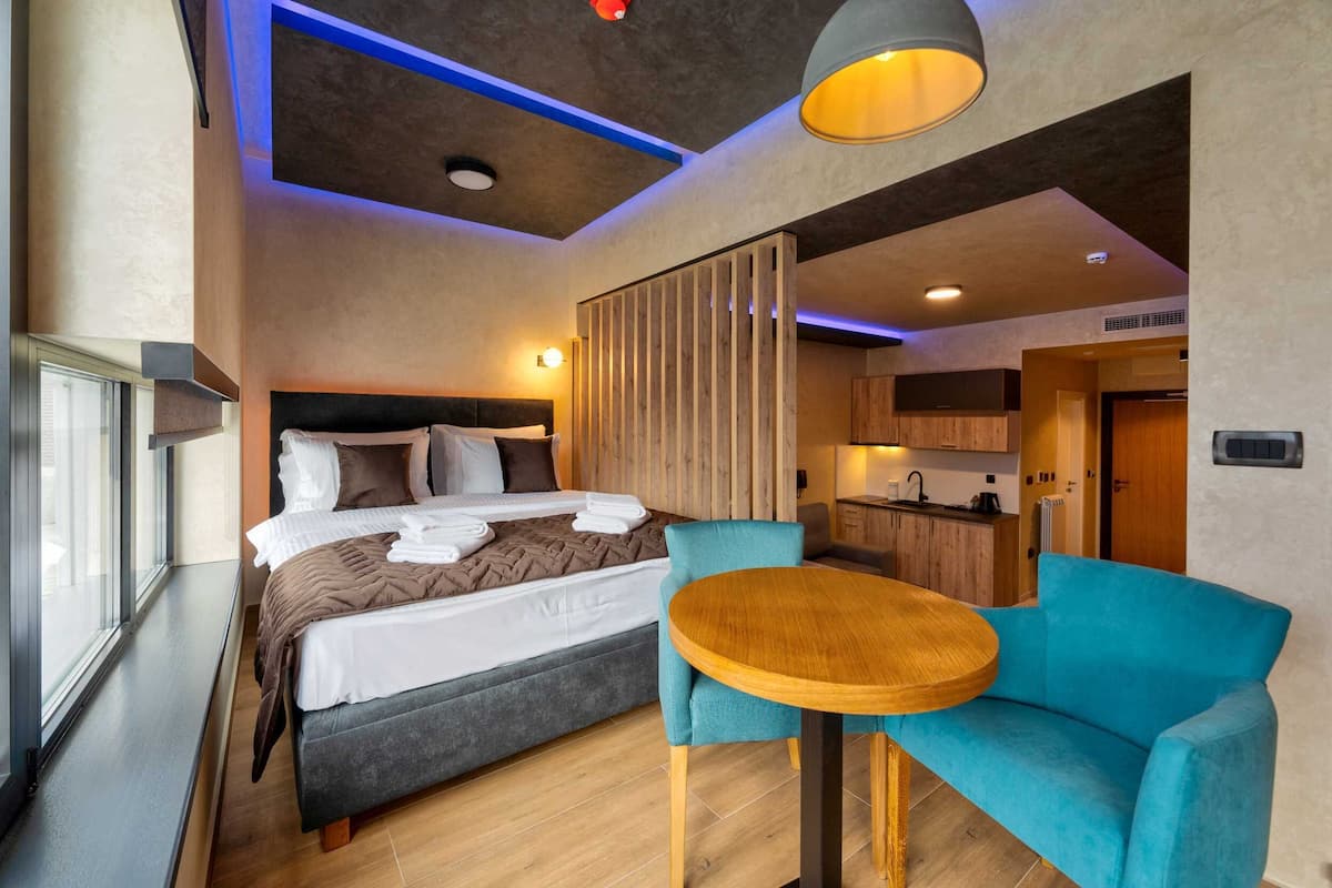 Habitación de hotel moderna con una cama grande, una mesita pequeña, un sillón azul y una kitchenette al fondo.