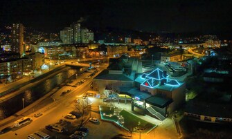 Vista aérea de un paisaje urbano nocturno con edificios iluminados, un río y un estacionamiento.