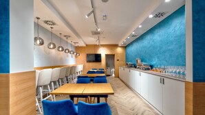 A modern restaurant with wooden tables and blue chairs, a kitchen area, and a blue accent wall. - Aria City Boutique Hotel (Užice)