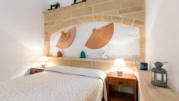 A bedroom with a bed, two bedside tables, a nightstand, a brick fireplace, and a shelf with decorative items.