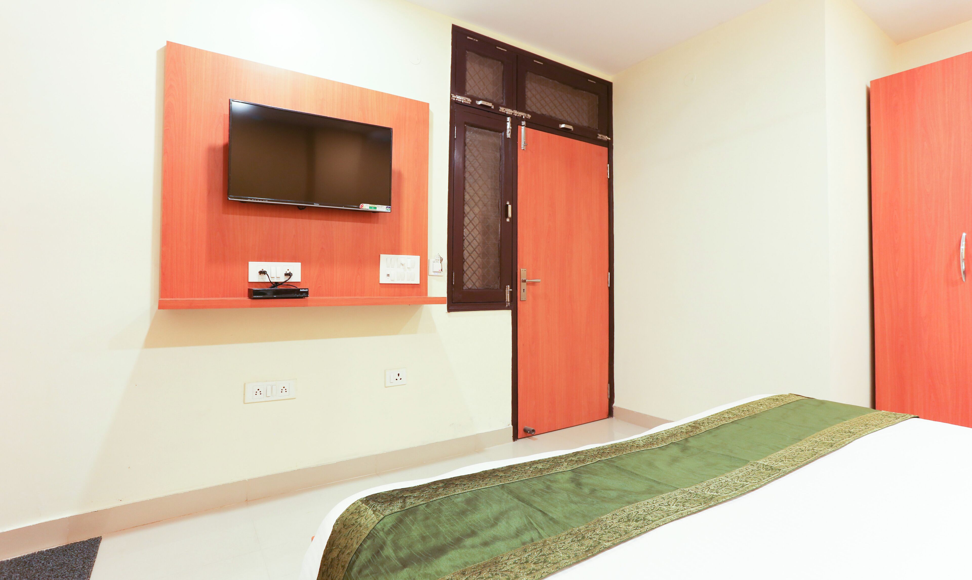A bedroom with a bed, a television mounted on the wall, a wardrobe, and a door with a window.