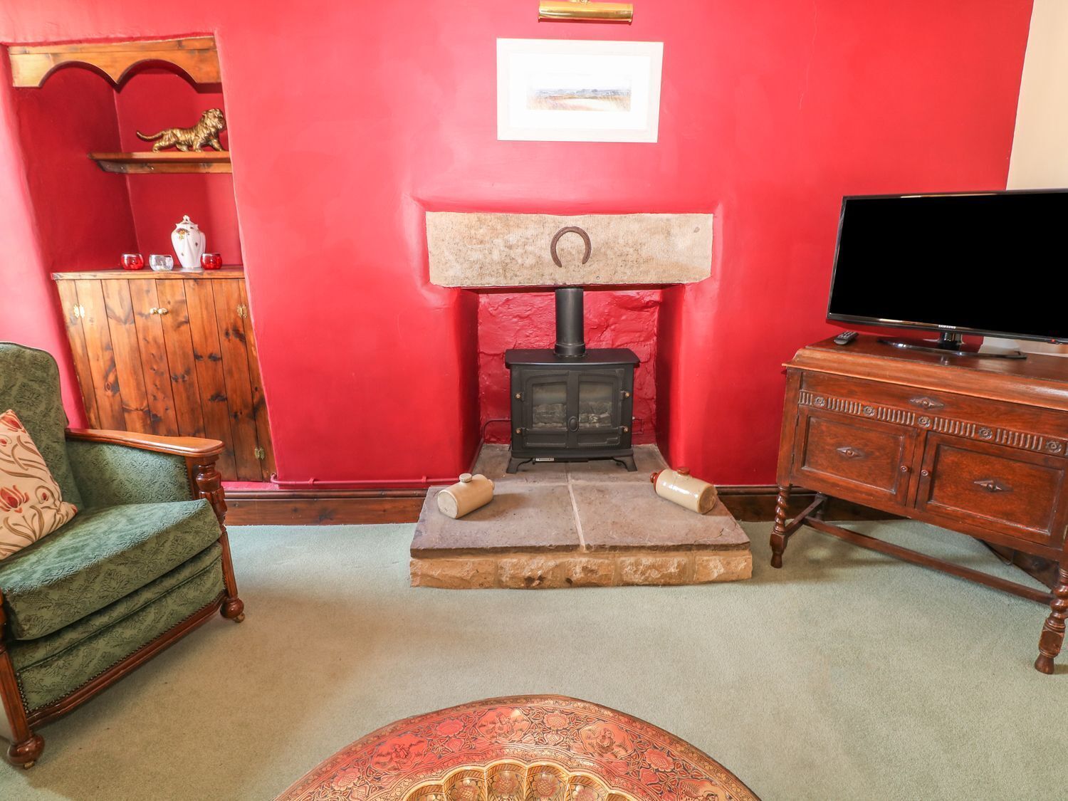 A living room with a red wall, a fireplace, a green armchair, a wooden cabinet, and a television.