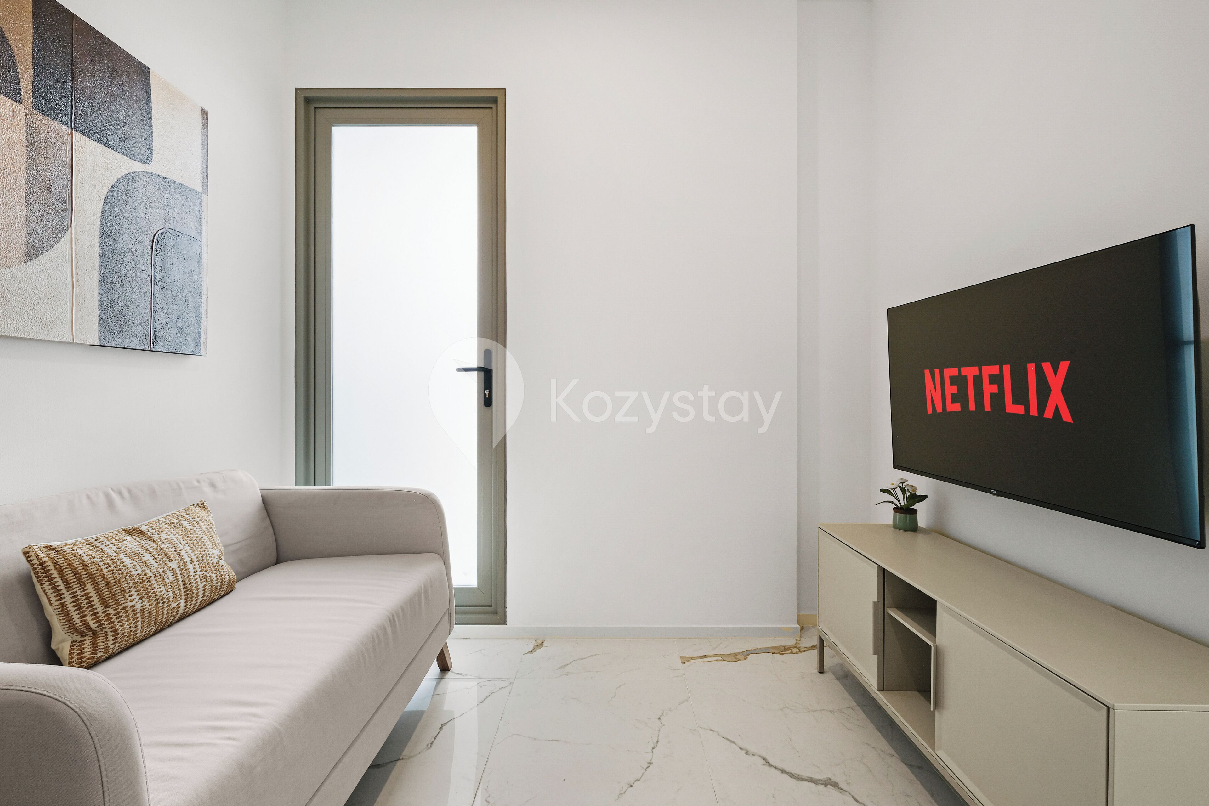 A modern living room with a beige sofa, a flat-screen TV mounted on the wall, and a minimalist decor.
