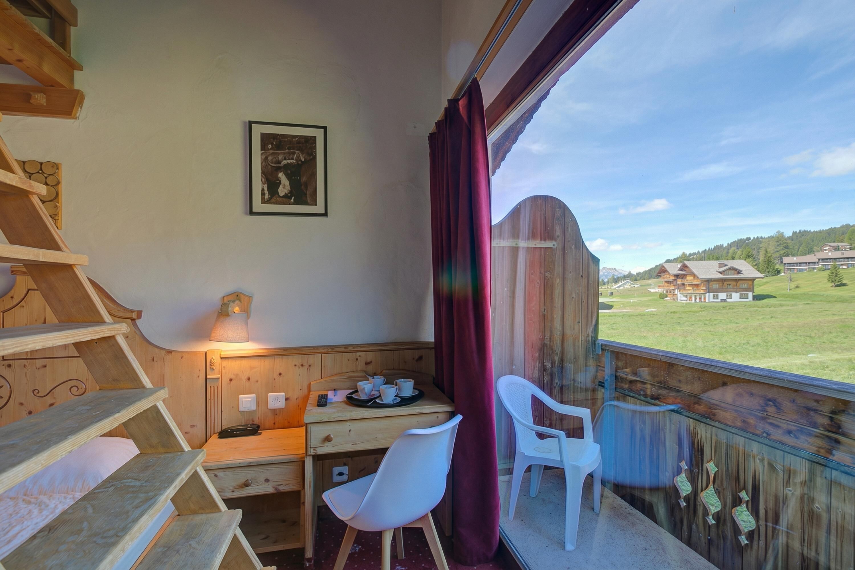 Ein Zimmer mit einem Holztisch, Stühlen, einem Bett, einem Fenster mit Blick auf Häuser und einem gerahmten Bild an der Wand.
