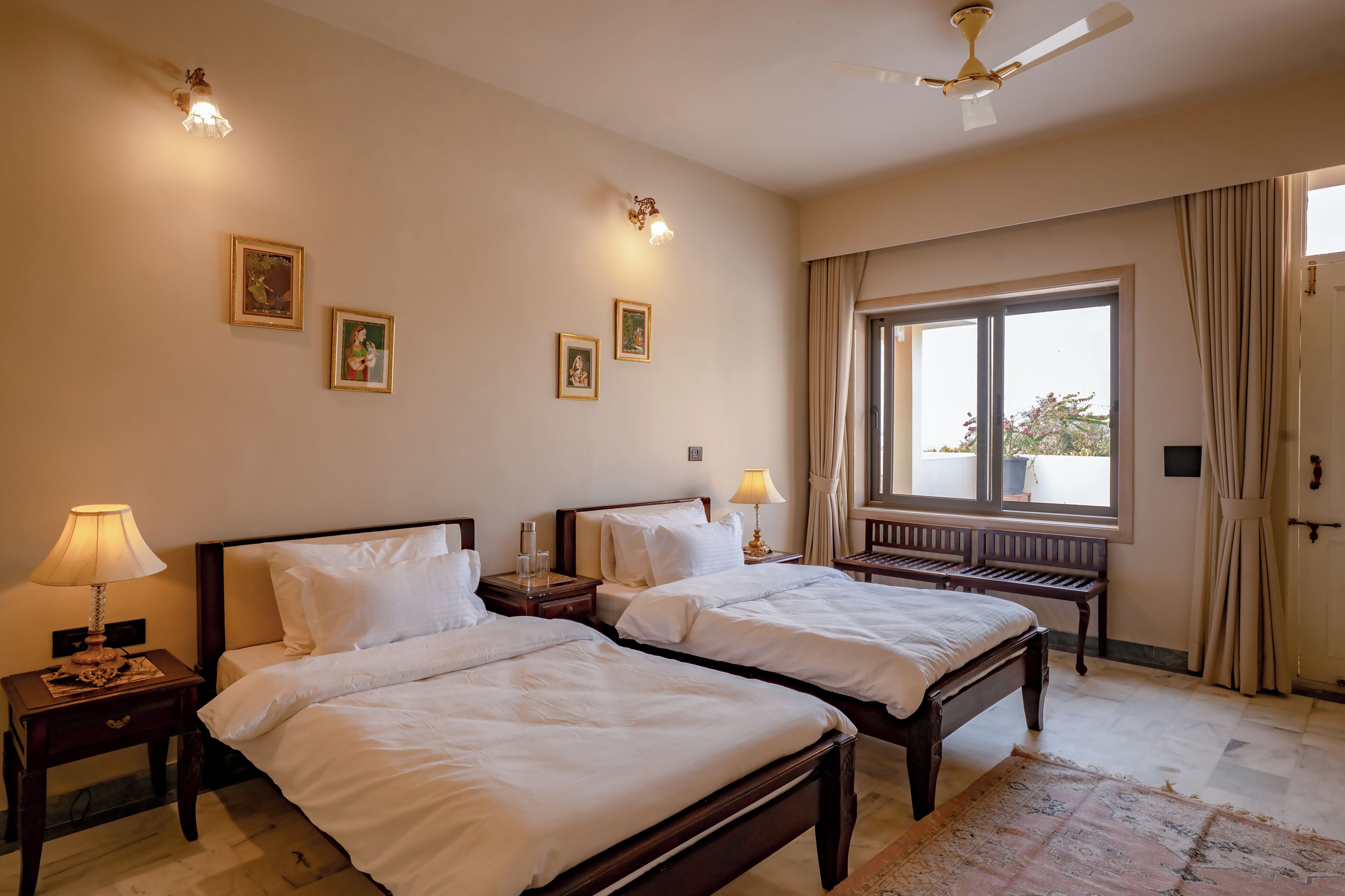 Habitación de hotel con dos camas, lámparas de noche, ventilador de techo y una ventana grande con cortinas.