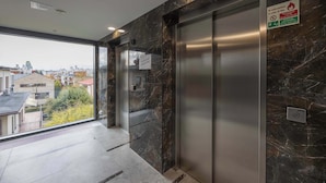 An elevator with a digital display, a fire safety sign, and a view of buildings through a glass door. - Functional Apartment | Large Balcony and Workspace (Warszawa)