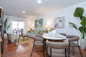 Uma sala de estar moderna com uma mesa de jantar redonda, cadeiras estofadas cinzas, um sofá com almofadas estampadas e uma planta grande em vaso.