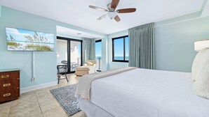 Un dormitorio con cama, ventilador de techo, televisor en la pared y vista al océano a través de la ventana.